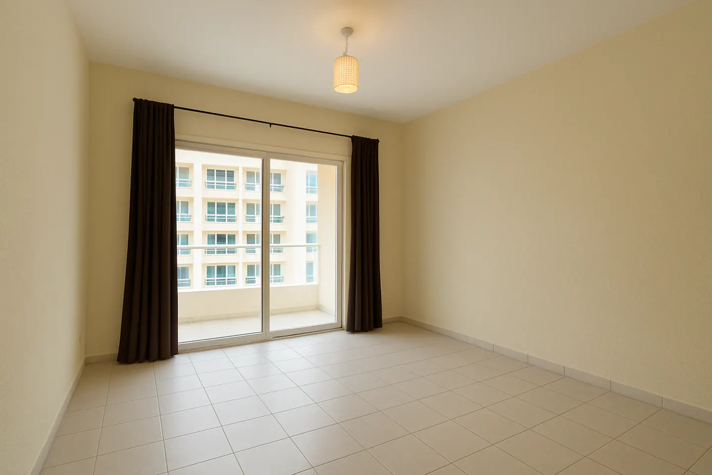 Empty room with beige walls, tile floor, and brown curtains framing a sliding glass door to a balcony. A light fixture hangs from the ceiling.