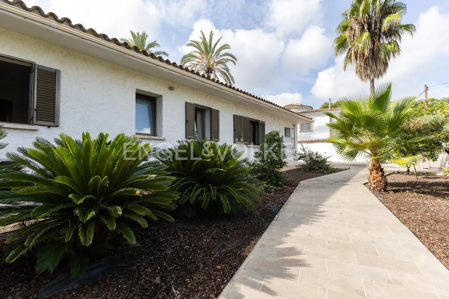 Exterior view of a white stucco house with brown shutters, a stone walkway, and lush green landscaping. Palm trees are visible in the background.