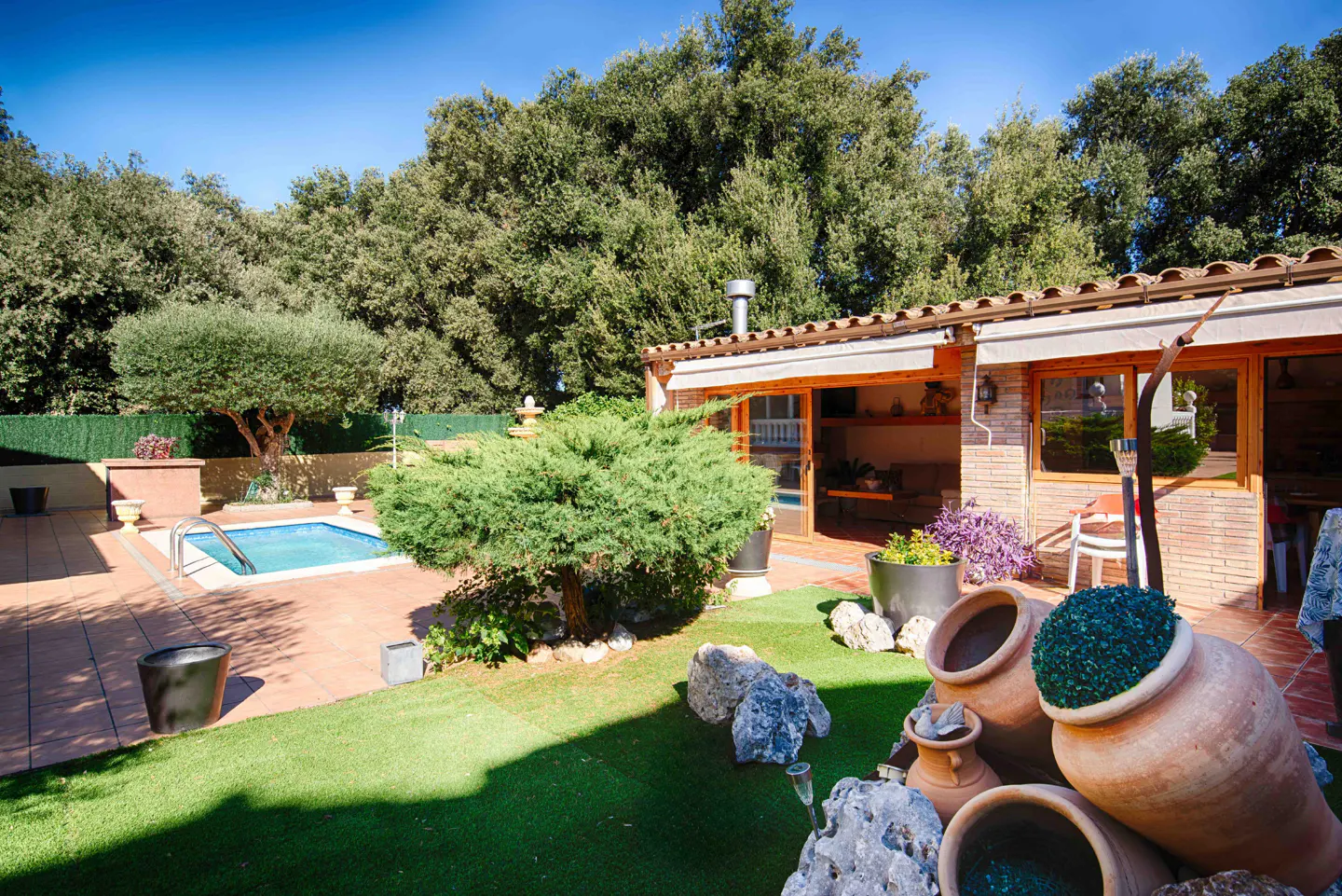 Backyard with a pool, brick building, and large clay pots on green grass. Trees in the background.