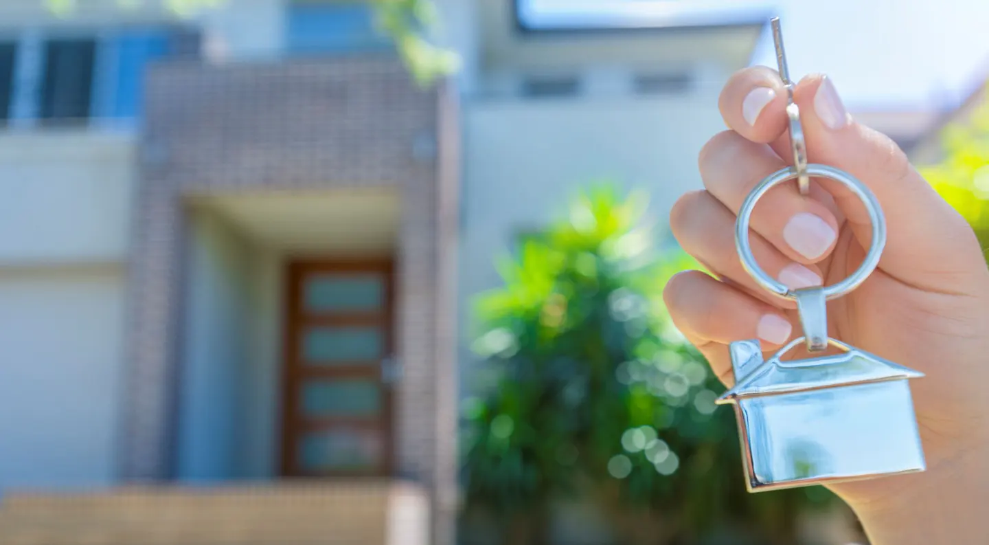 A hand holds keys with a house-shaped keychain in front of a blurred house.