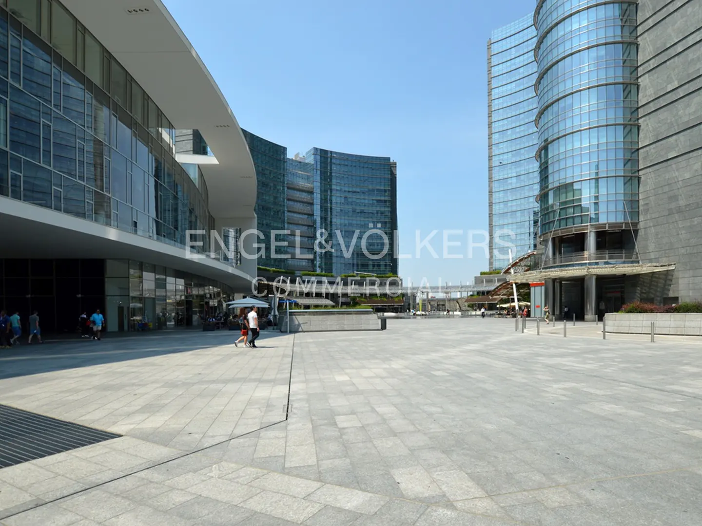Modern city square with glass skyscrapers and people walking. The Engel & Völkers logo is visible.