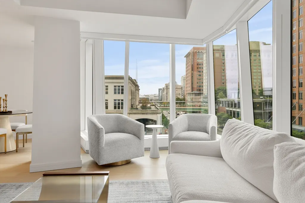 Bright living room with white sofa, two gray chairs, and a view of city buildings through large windows.