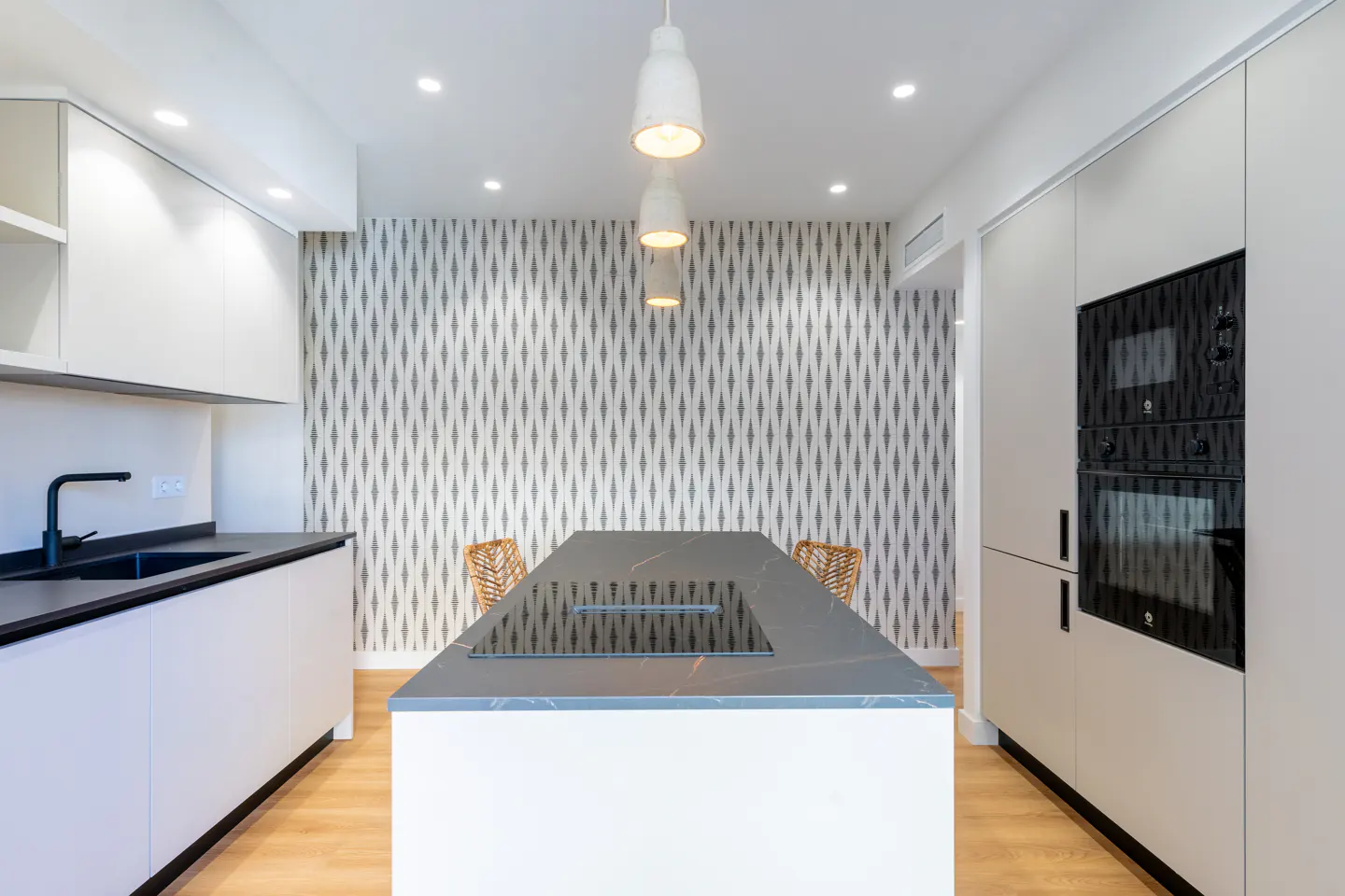 Bright kitchen with white cabinets, black countertops, and a gray island with a cooktop. Patterned wallpaper and wood floors add texture.