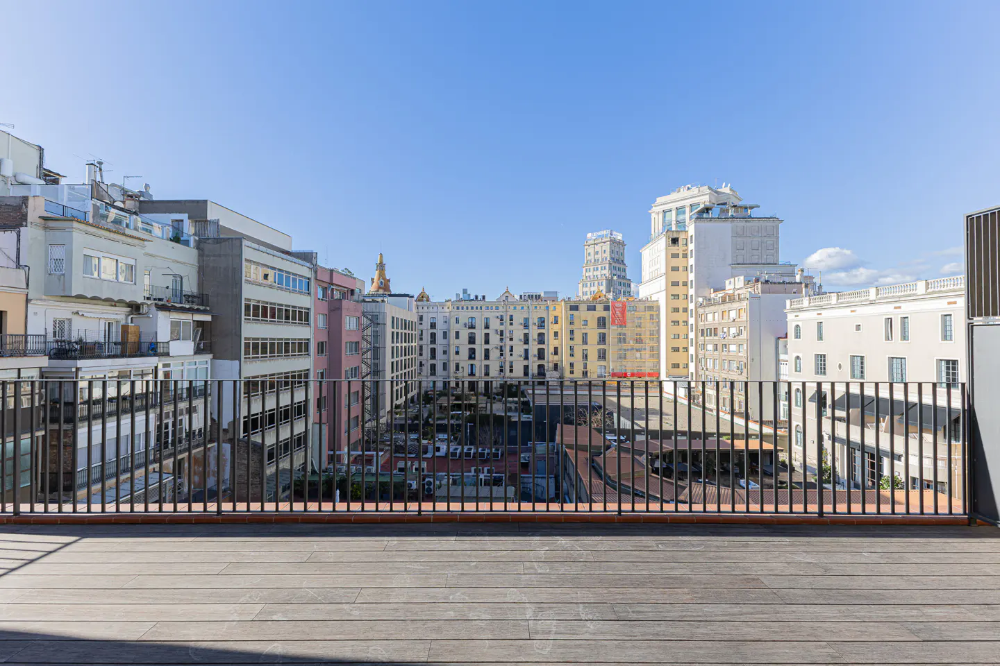 View from a wooden rooftop terrace with a black metal railing overlooking city buildings under a clear blue sky.