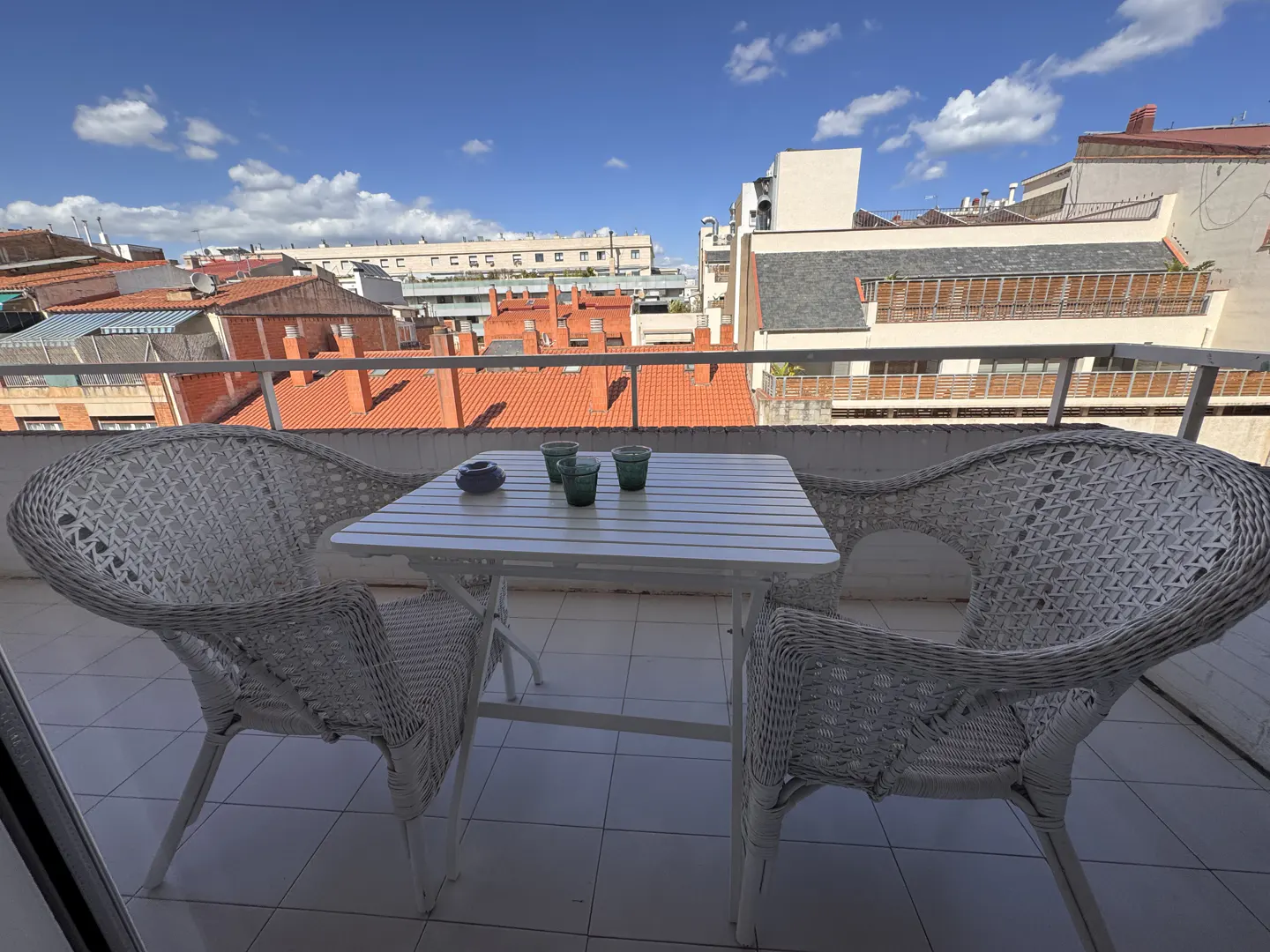 Balcony view with white wicker chairs and table set with glasses, overlooking red-roofed buildings under a blue sky.