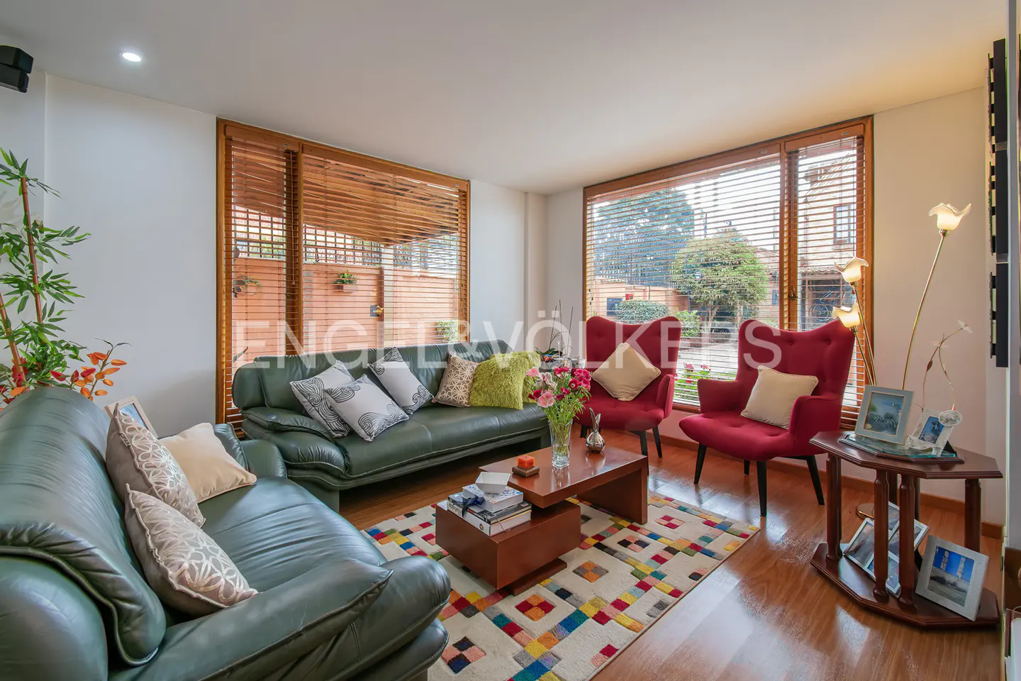 Living room with green leather sofas, red chairs, wood floors, and a colorful rug. Windows with blinds overlook a garden.