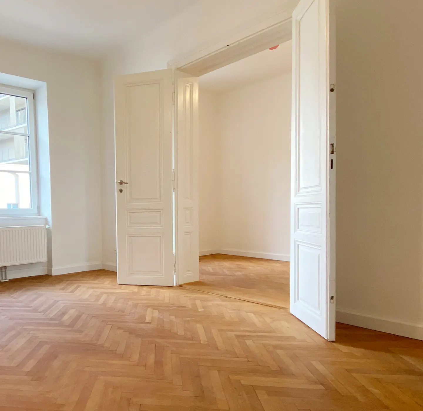 Bright, empty room with herringbone wood floors, white walls, and open white double doors leading to another room. A window is on the left.
