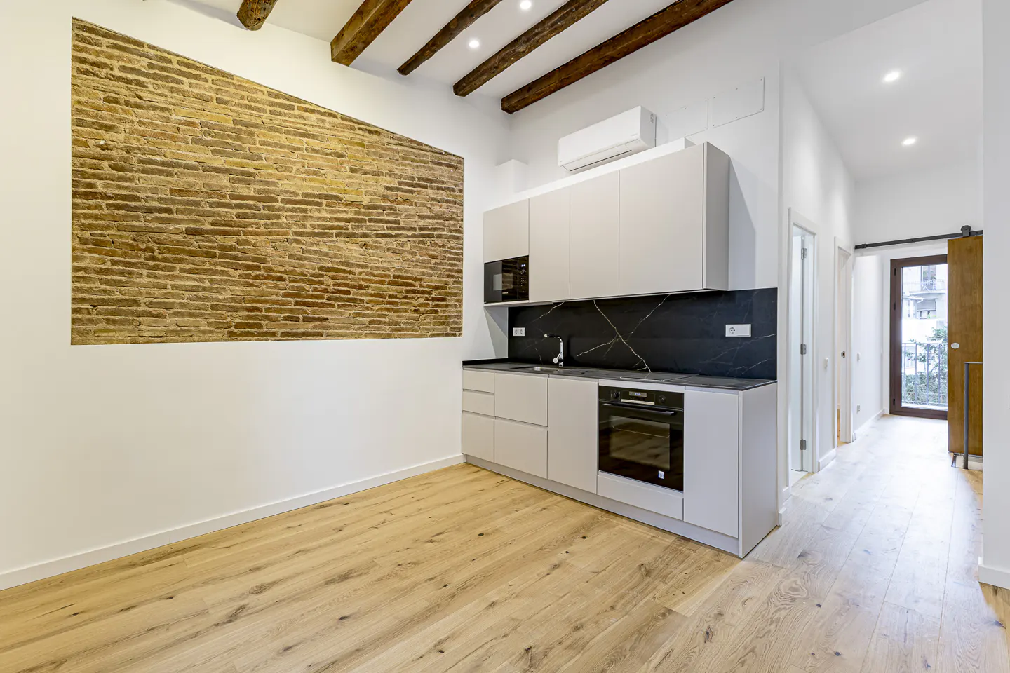 Modern kitchen with light wood floors, exposed ceiling beams, a brick accent wall, and sleek white cabinets.
