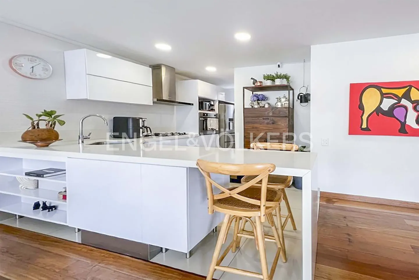 A modern kitchen with white cabinets, a white countertop, and two wooden bar stools. A colorful painting hangs on the wall.