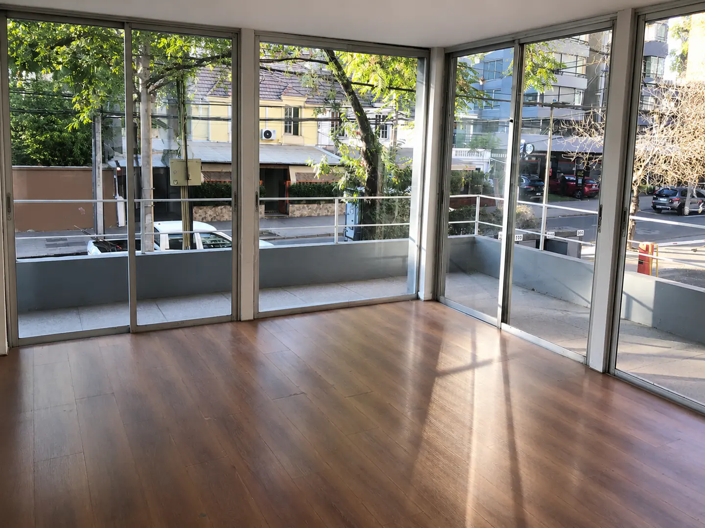 Empty room with wood floors and large sliding glass doors to a balcony overlooking a street with trees and buildings.