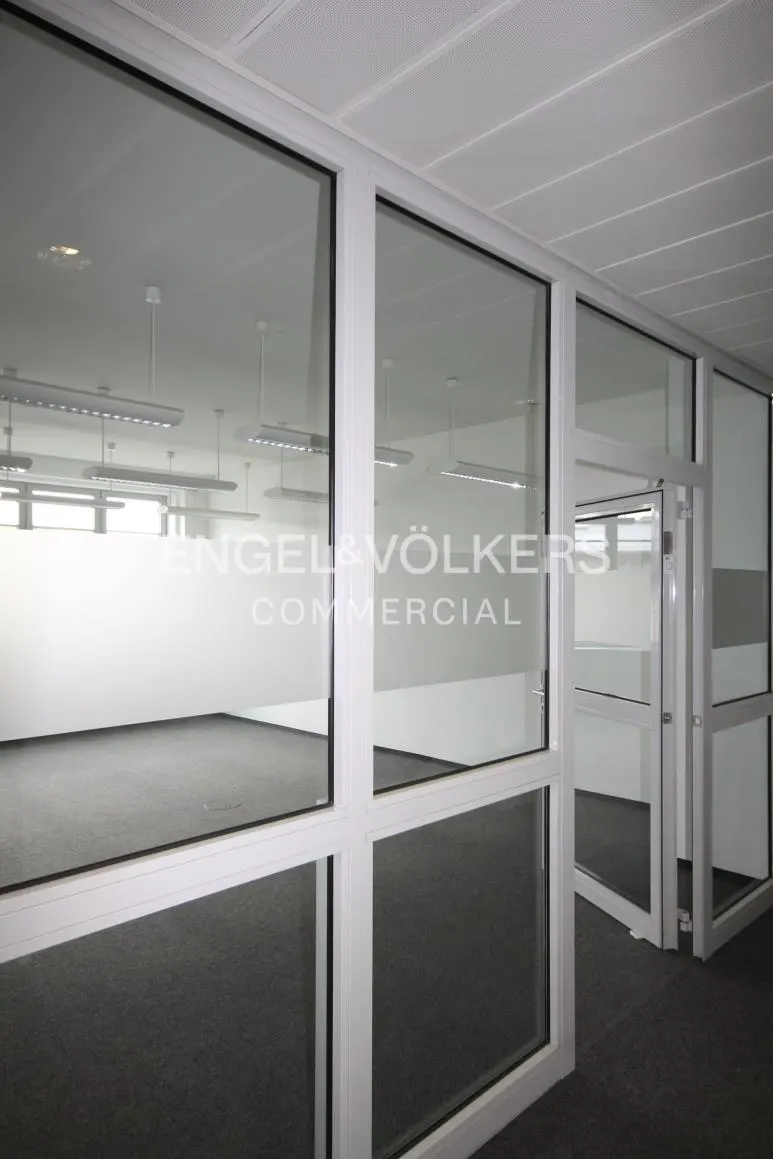 Interior view of a commercial space with white walls, gray carpet, and glass-paneled partitions. The Engel & Völkers Commercial logo is visible on the glass.