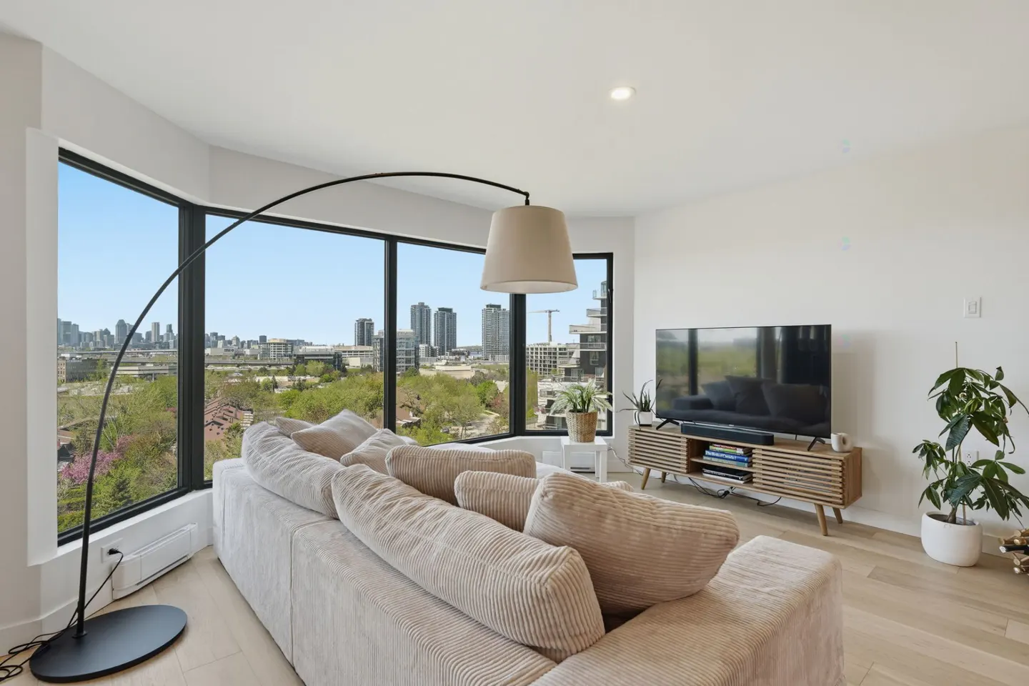 Bright living room with a beige corduroy sofa, black arc lamp, and a large window overlooking a city skyline. A TV sits on a wooden console table.