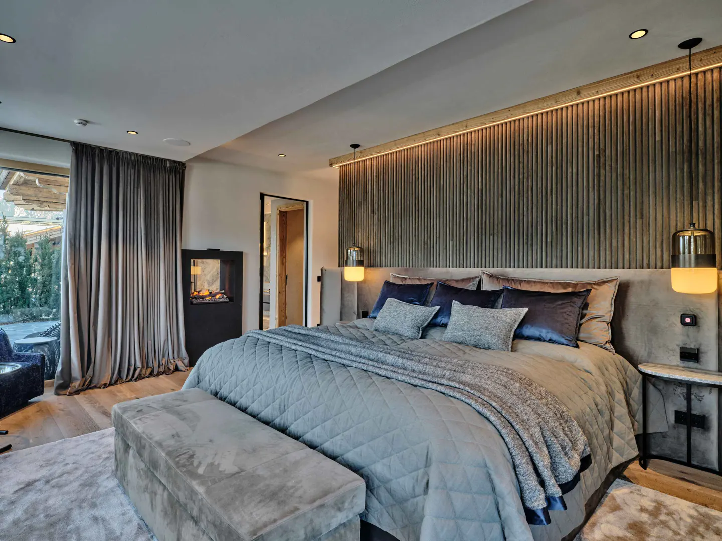 A modern bedroom with a large bed, gray bedding, and a wooden headboard. A fireplace is on the left, and a window with curtains shows an outdoor view.