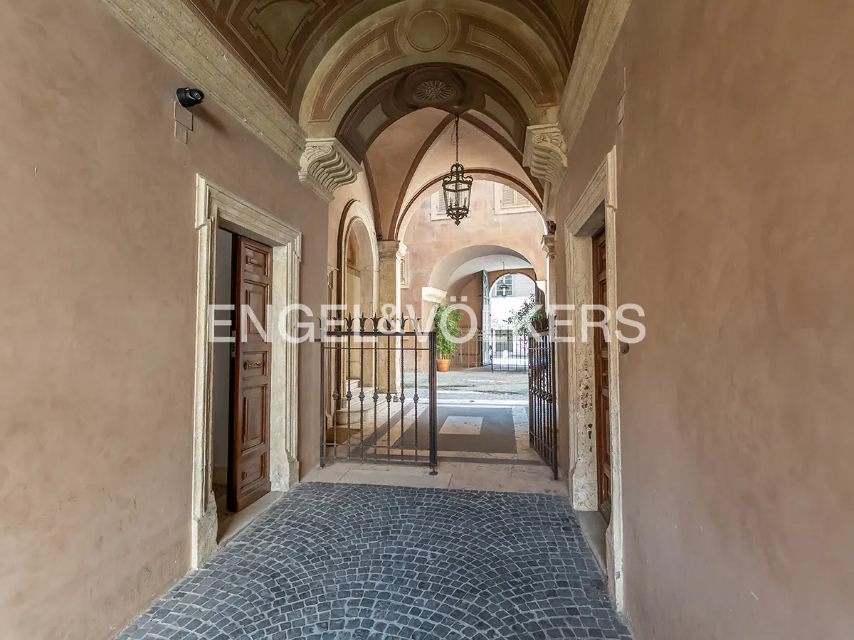 Arched walkway with stone floors and walls. A black metal gate leads to an open courtyard with a hanging lantern.