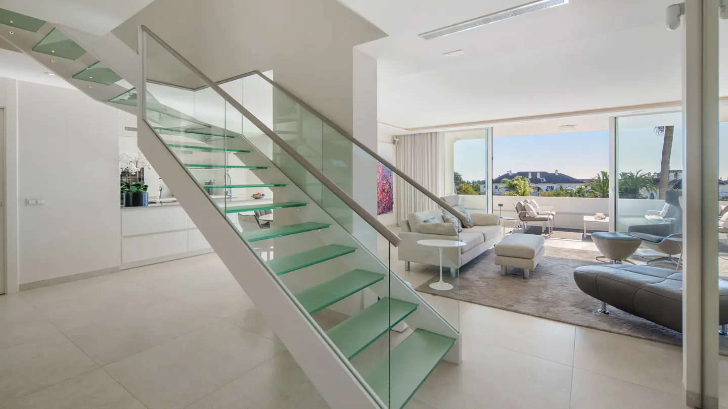 Bright, modern interior with glass staircase, white walls, and open living area. Balcony with outdoor seating visible through sliding glass doors.
