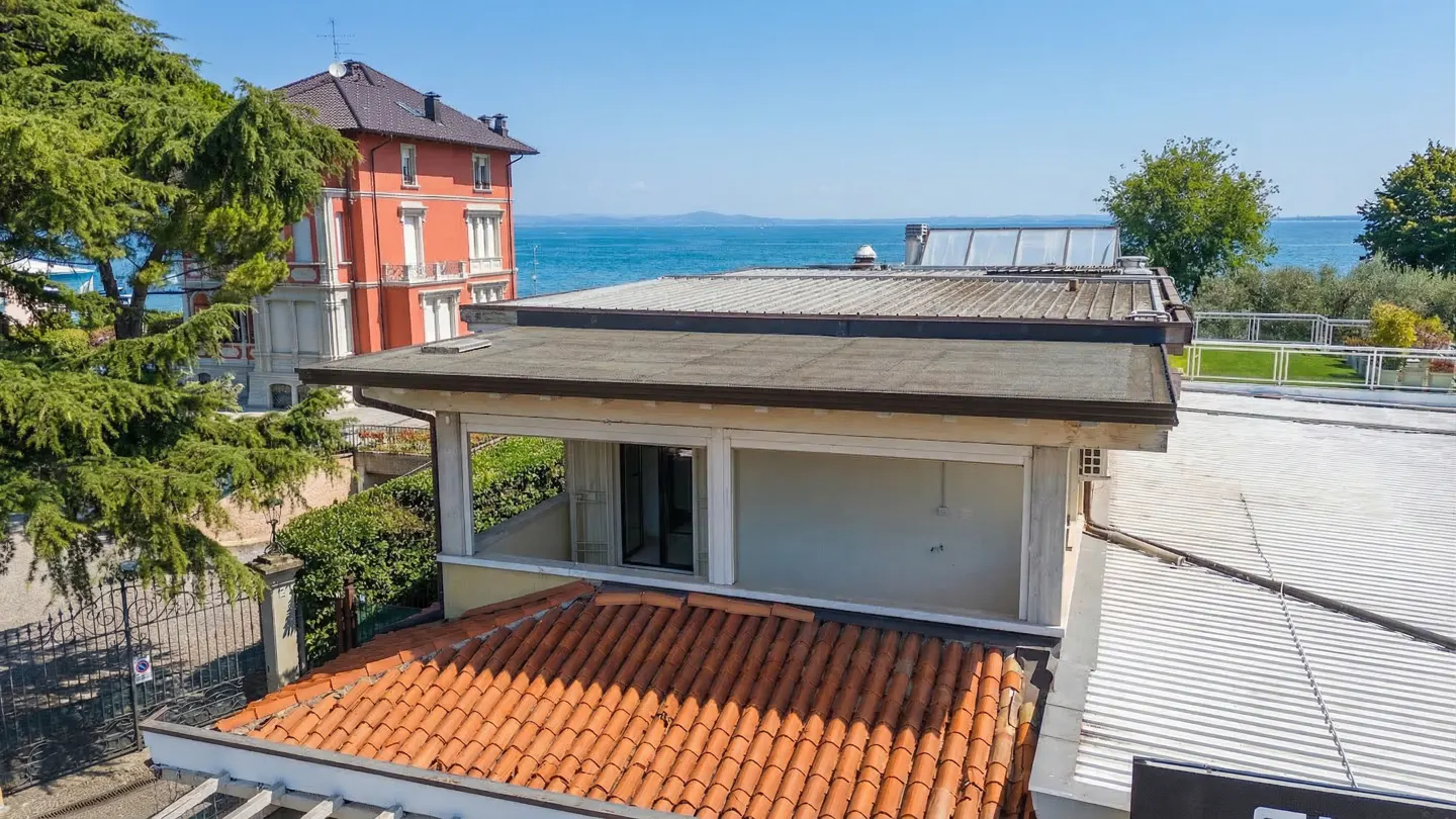 A high-angle shot shows rooftops with orange tiles, a balcony, and a view of a lake and a red building in the background.