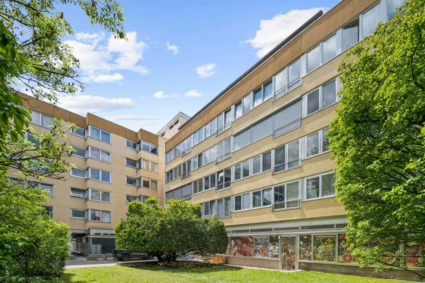 Exterior view of a multi-story tan apartment building with large windows, surrounded by green trees and a grassy courtyard.