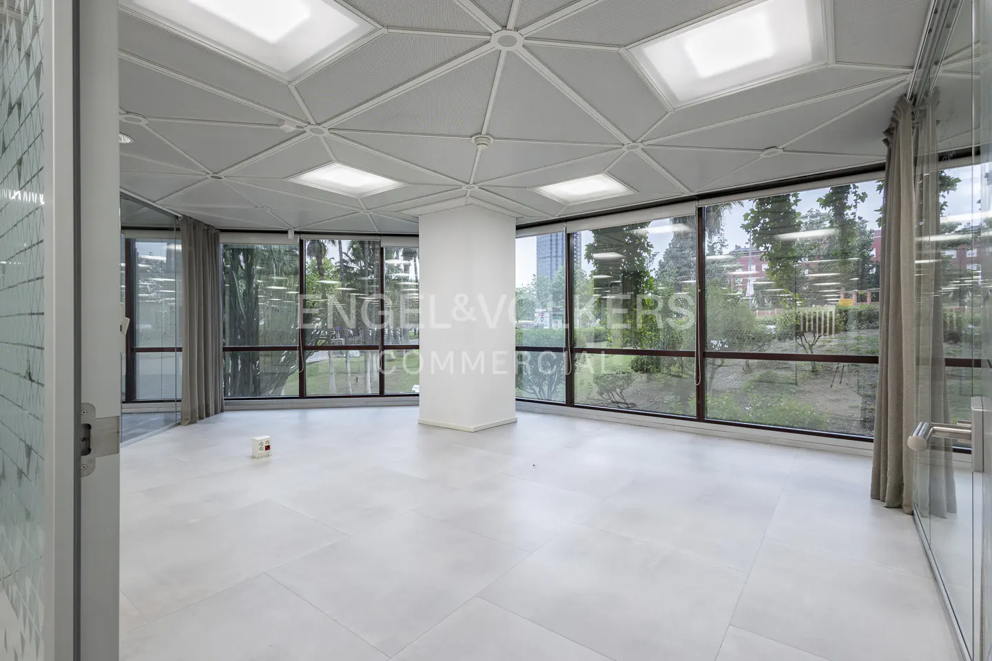 Bright, empty office space with white tile floors, large windows, and a geometric ceiling. Outside, greenery is visible.