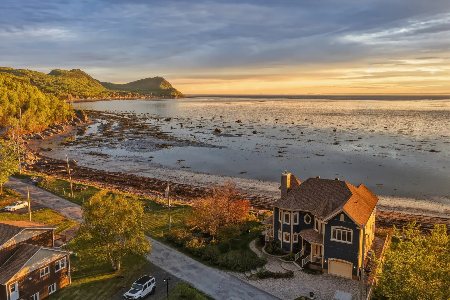 Aerial view of a blue two-story house by a rocky coastline at sunset, with mountains in the distance and a paved driveway.
