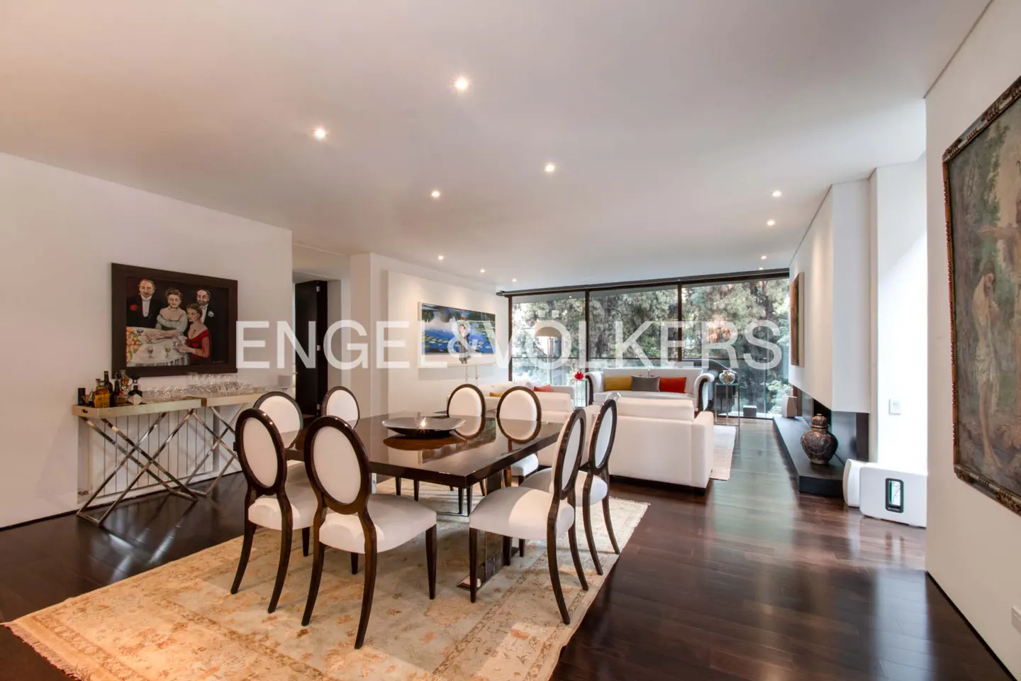 Open-concept living and dining area with dark wood floors, white walls, and large windows overlooking trees. Dining table with white chairs.