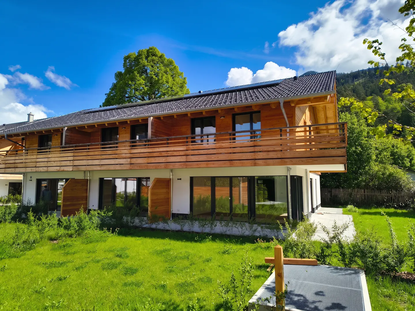 Two-story house with wood siding and a balcony, set against a blue sky with green lawn.
