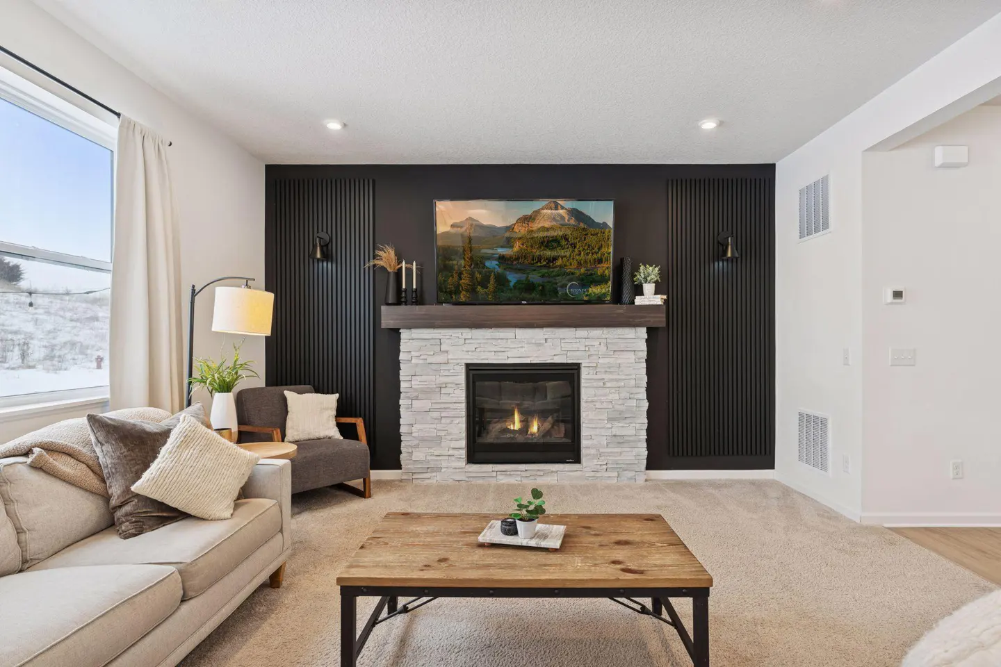 Living room with a stone fireplace, dark wood mantle, and TV. A beige sofa and wood coffee table sit on a light carpet.