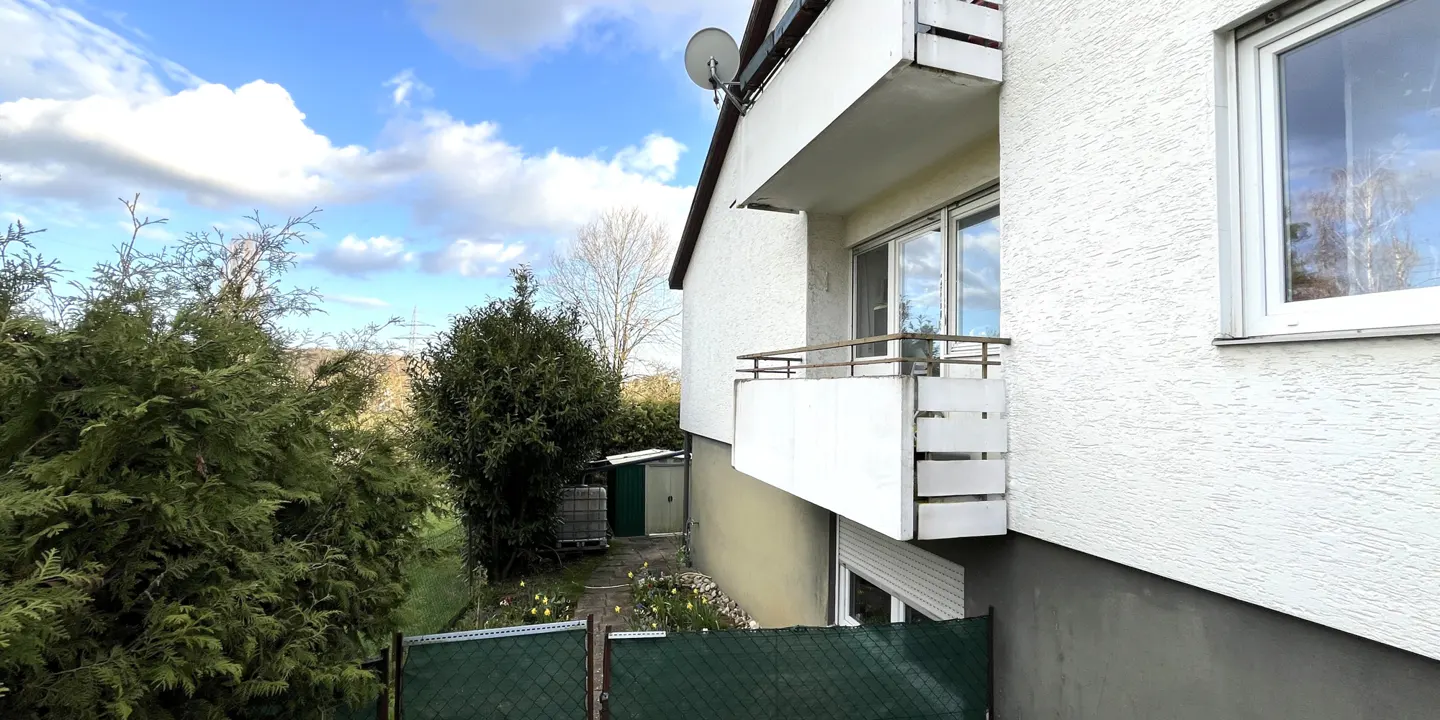 Exterior view of a white building with balconies, green trees, and a blue sky with clouds.
