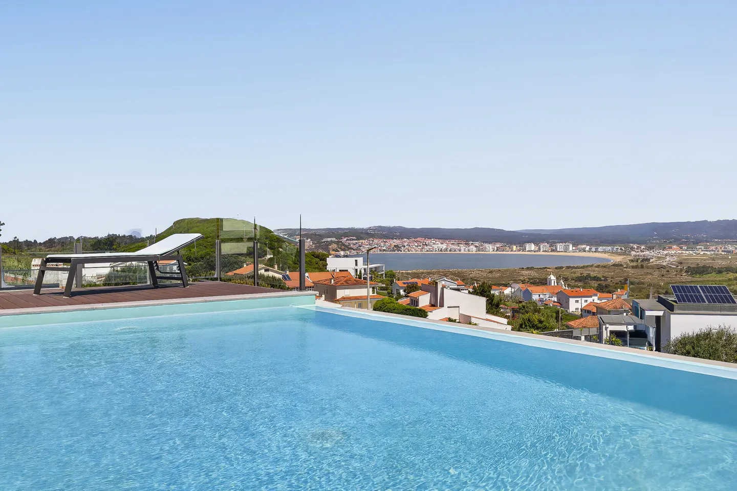 Rooftop pool with blue water, lounge chair, and a view of a town and lake under a clear blue sky.