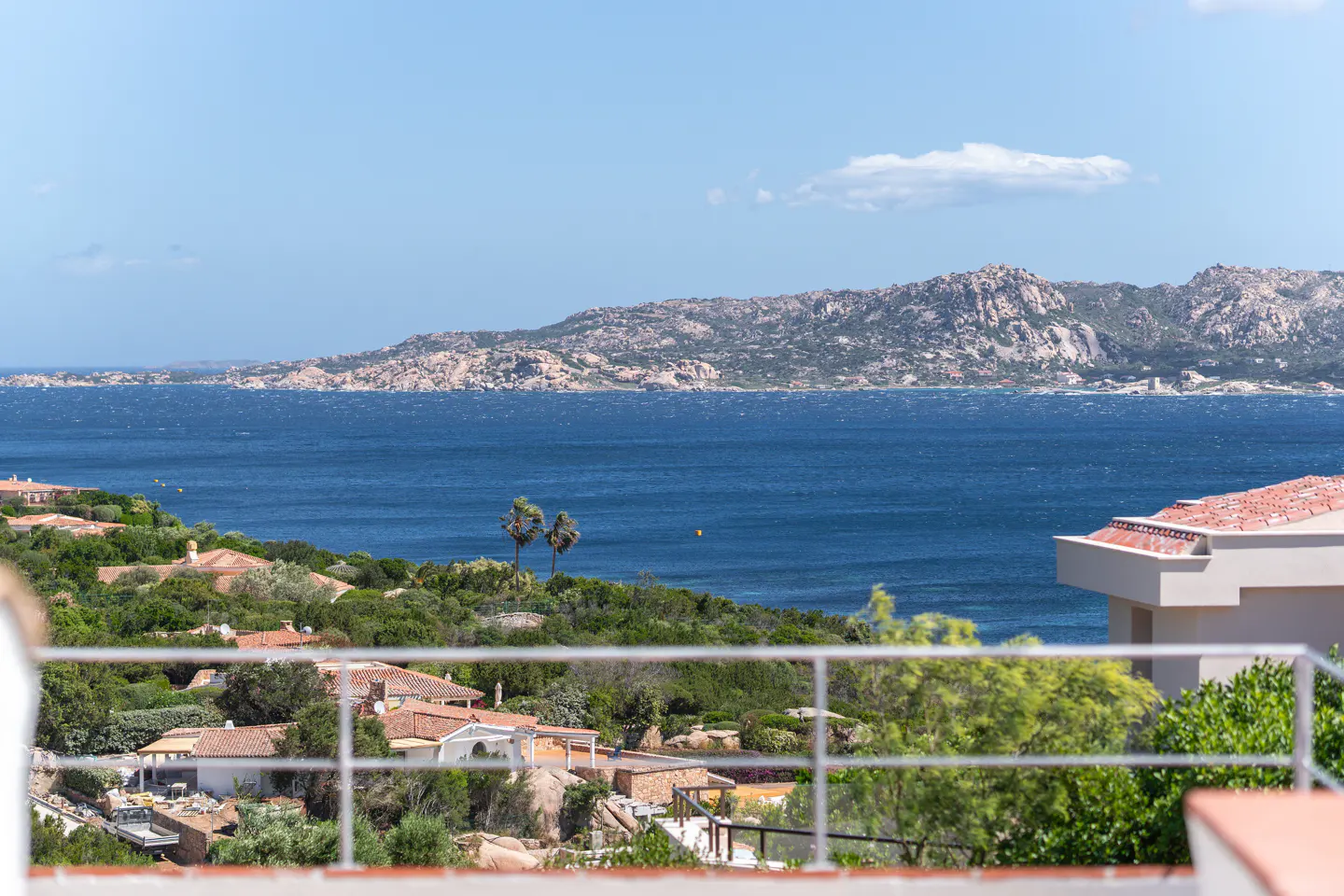 View from a balcony overlooking blue ocean water, green trees, and houses with red tile roofs. A rocky island is in the distance.