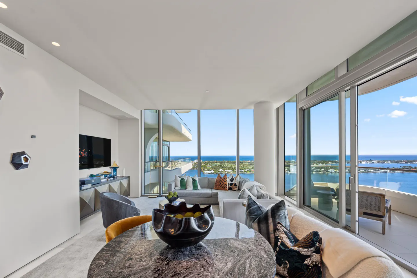 Bright, modern living room with floor-to-ceiling windows overlooking the ocean. A marble table and sectional sofa furnish the space.