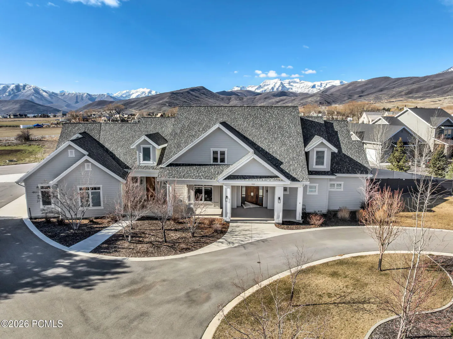 Aerial view of a light gray house with a dark gray roof, a circular driveway, and snow-capped mountains in the background.