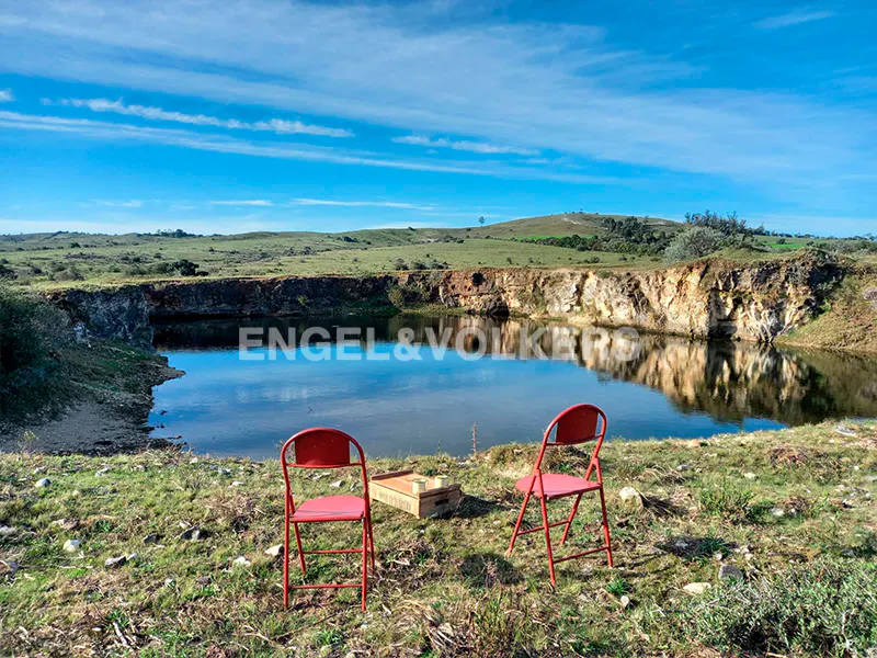 Two red chairs face a calm pond set within a rocky landscape under a bright blue sky.