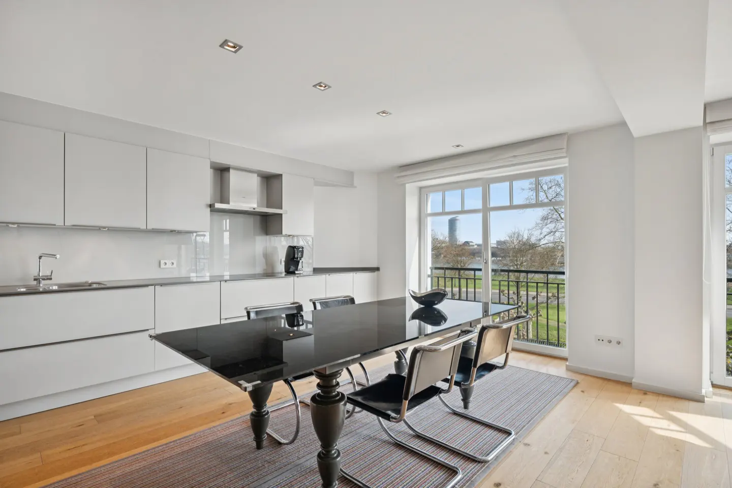 Bright, modern kitchen with white cabinets, a black dining table, and a view of a park and city skyline through a large window.