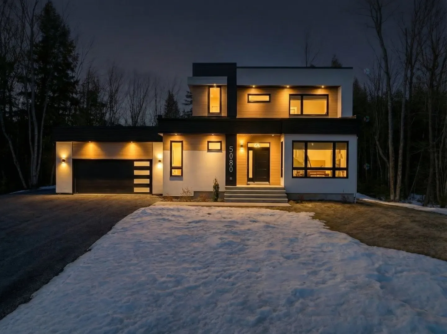 Two-story modern home with a black garage door and snow on the lawn at dusk.