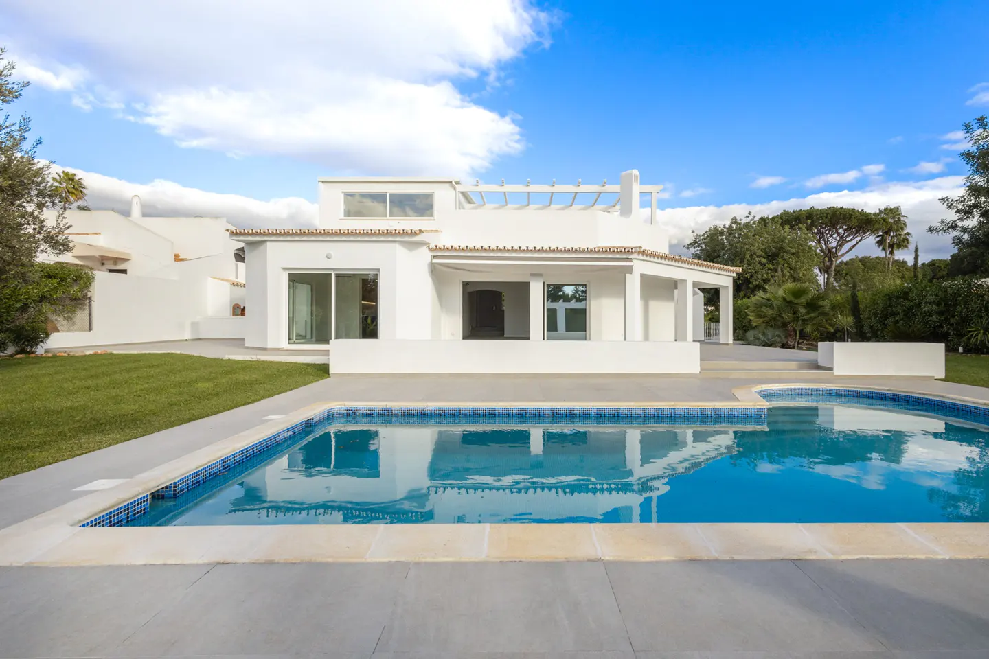 Modern white house with a blue tiled pool, green lawn, and blue sky with white clouds.