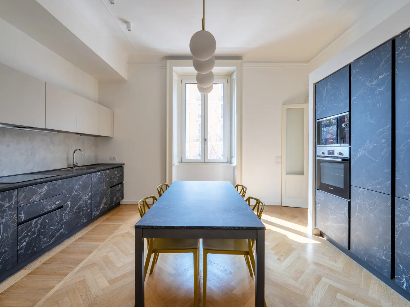 Bright kitchen with marble cabinets, white walls, and herringbone wood floors. A long table with yellow chairs sits under a globe pendant light.