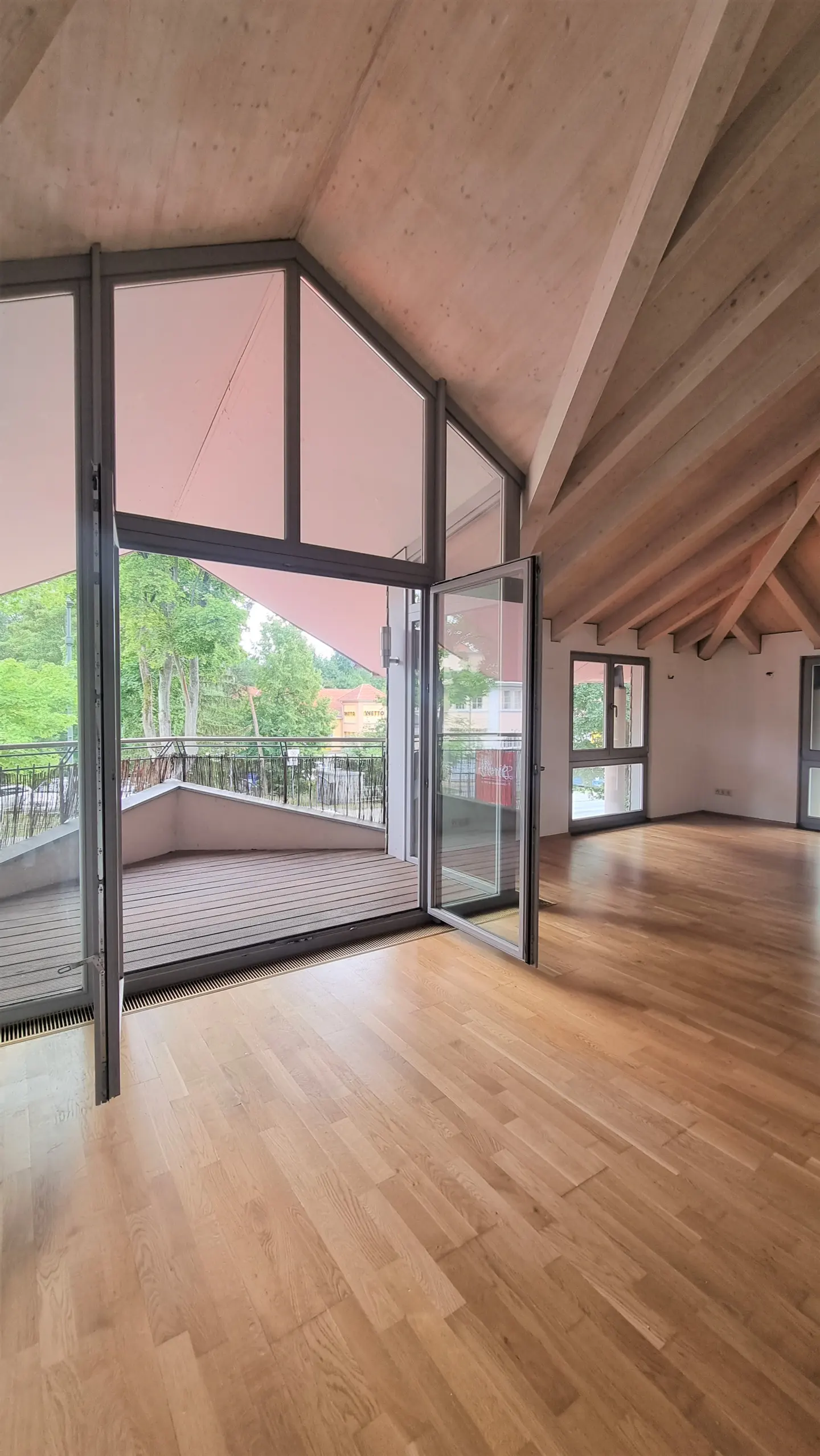 Bright, empty attic room with wood floors, exposed beams, and large windows opening to a balcony with a view of trees.