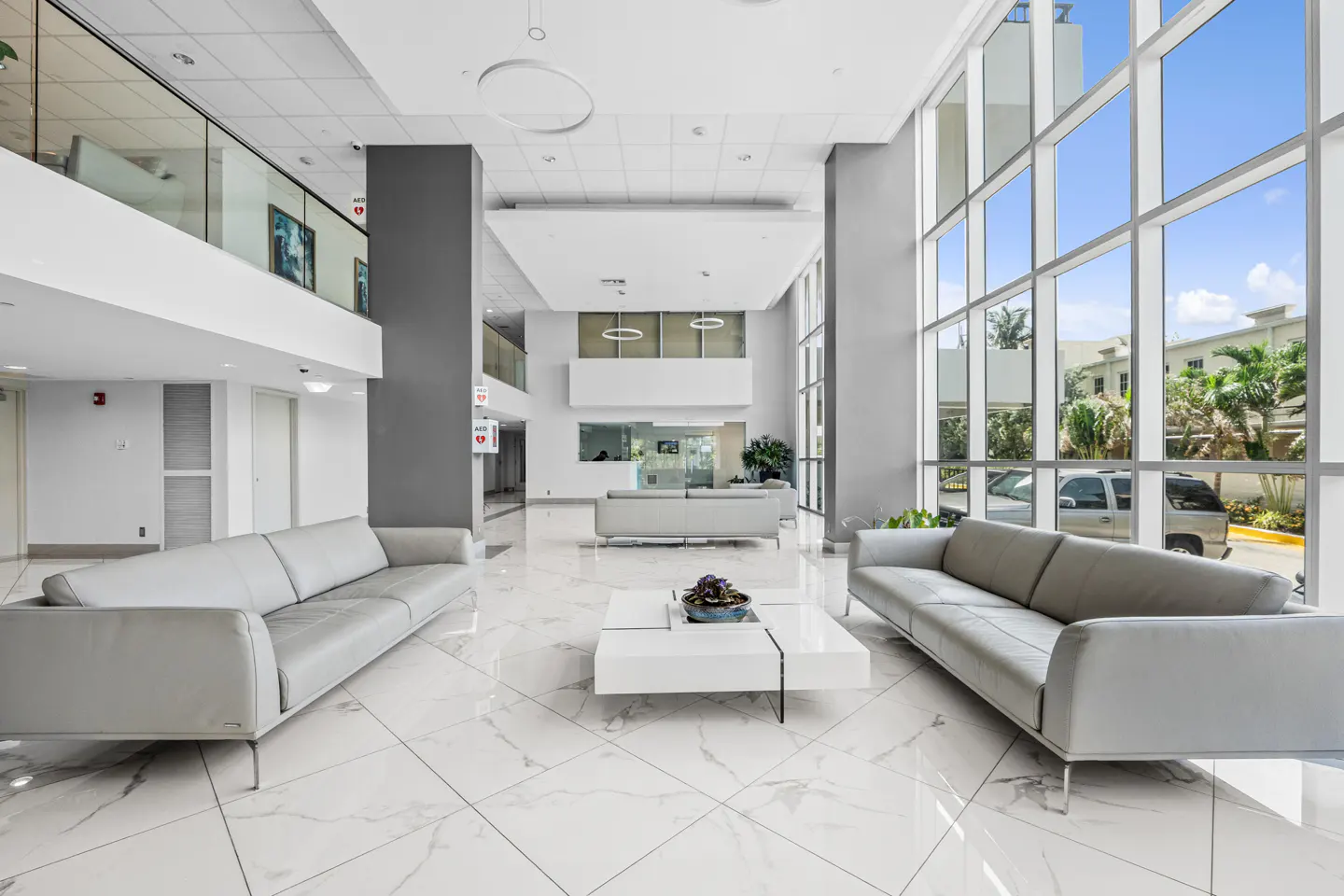 Bright, modern lobby with gray sofas, white marble floors, and floor-to-ceiling windows showing palm trees.
