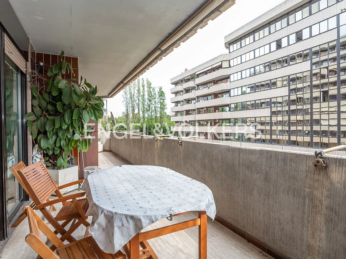 Balcony view with wooden table, chairs, and a large green plant. A tall building is visible in the background.