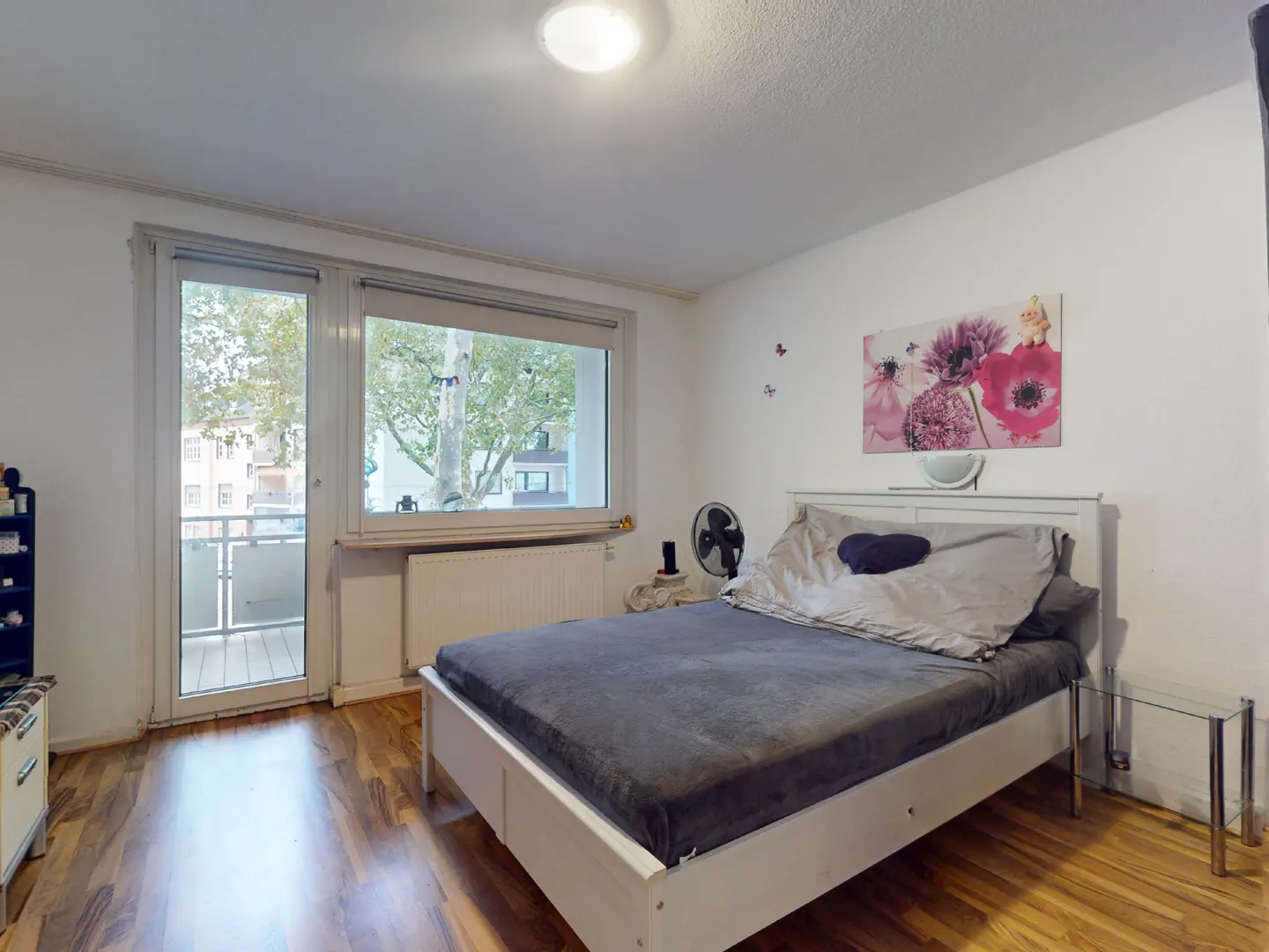 Bedroom with a white bed frame, gray bedding, wood floors, and a balcony door. A flower painting hangs on the wall.