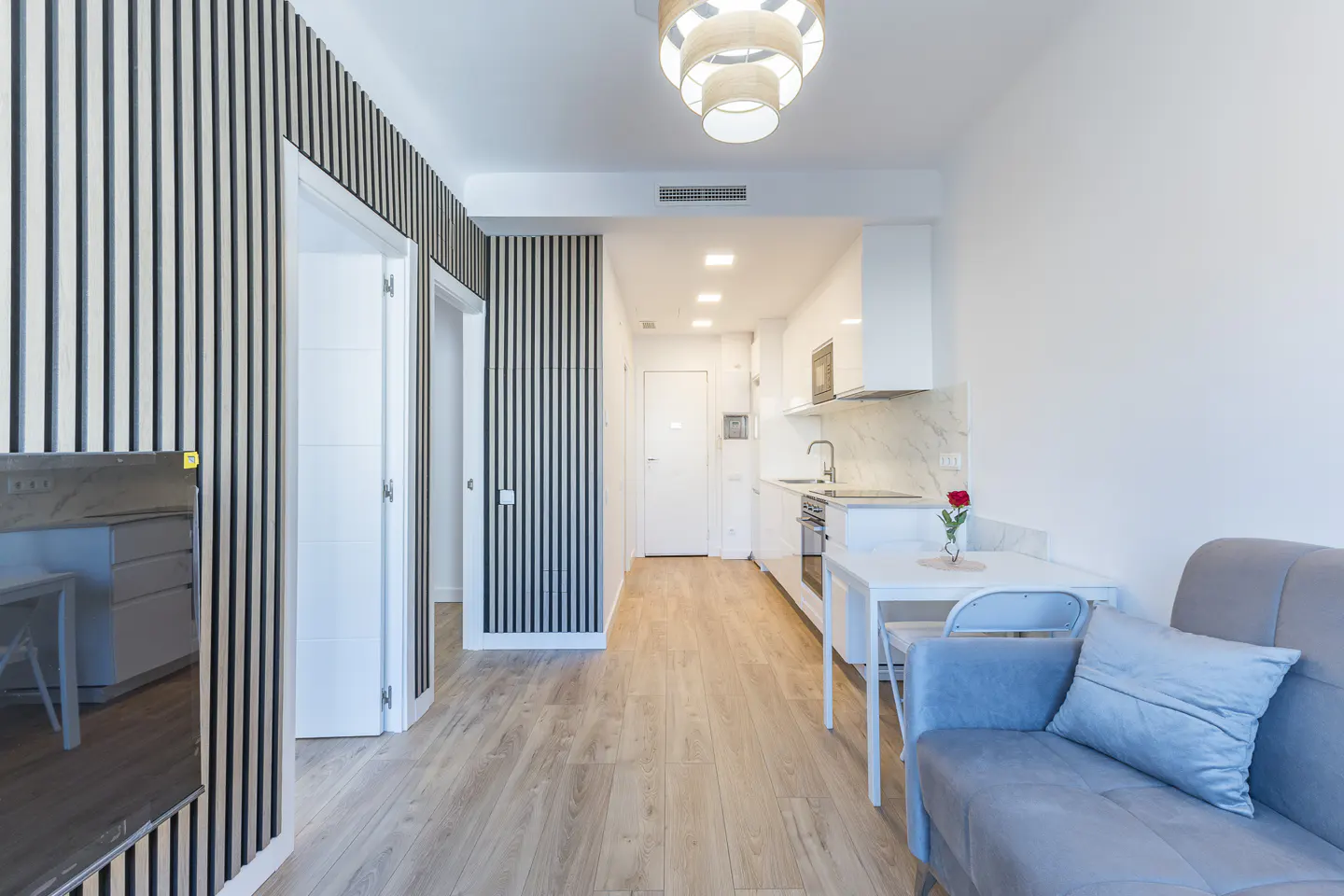 Modern studio apartment with wood-slat accent walls, light wood floors, a grey sofa, and a compact white kitchen.