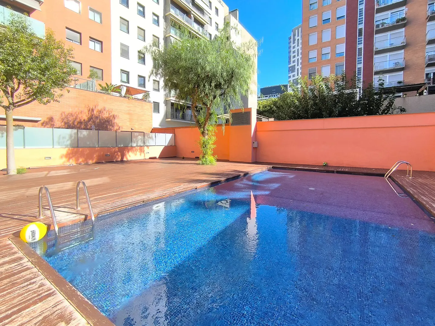 Outdoor pool with blue tiles and wooden deck, surrounded by peach-colored walls and apartment buildings under a clear blue sky.