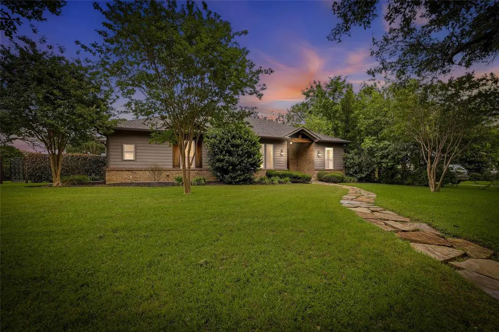 A single-story home with gray siding, brick accents, and a stone walkway leading to the front door. Green lawn and trees surround the house.
