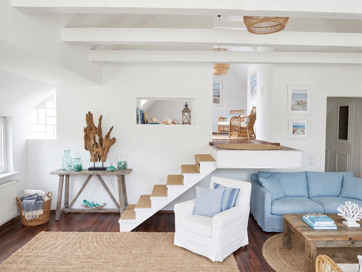Bright living room with white walls, wood floors, and a blue sofa. A wooden table displays glass vases and driftwood art. Stairs lead to an upper level.