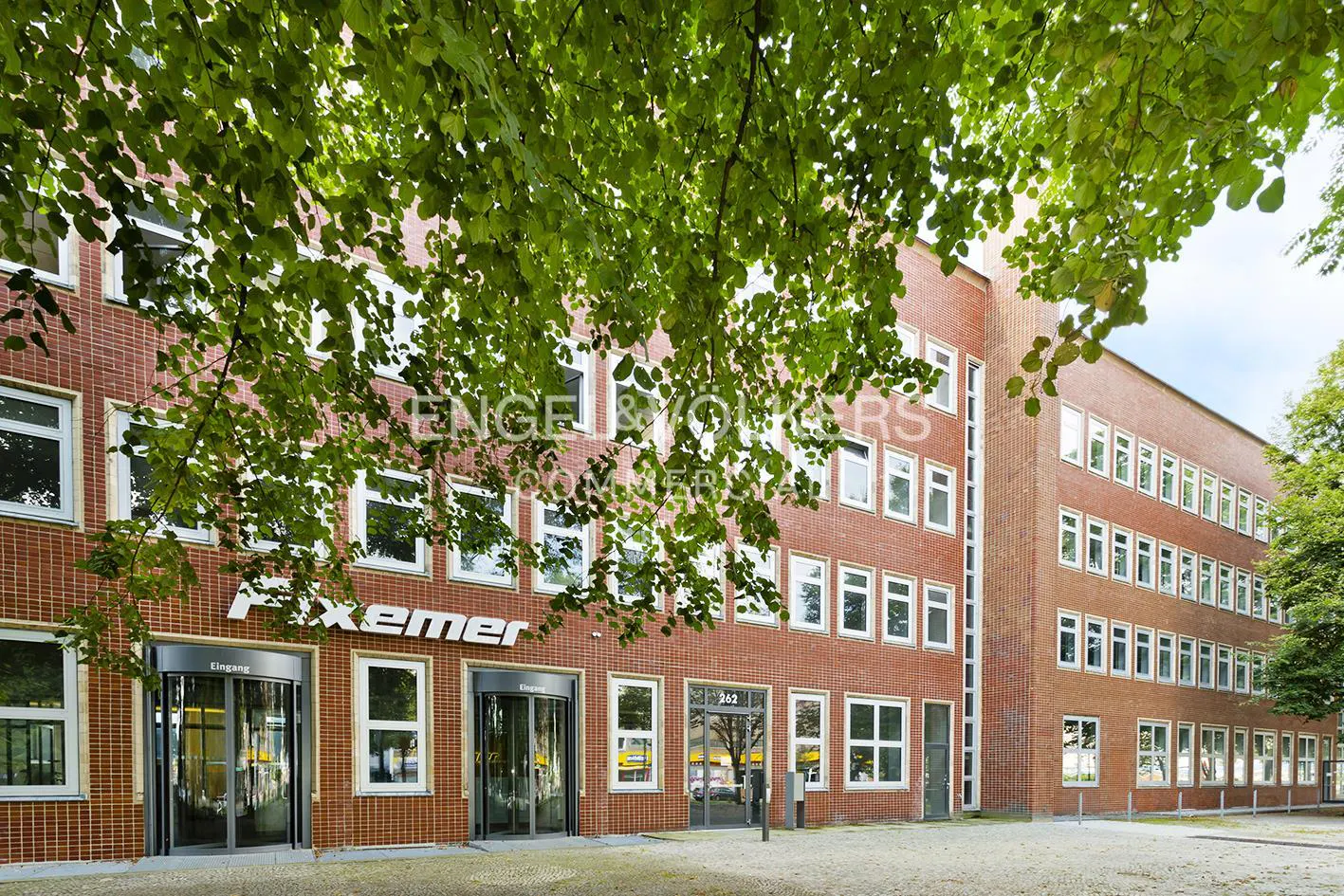 Exterior view of a red brick building with white framed windows and revolving doors. Green leaves are in the foreground.