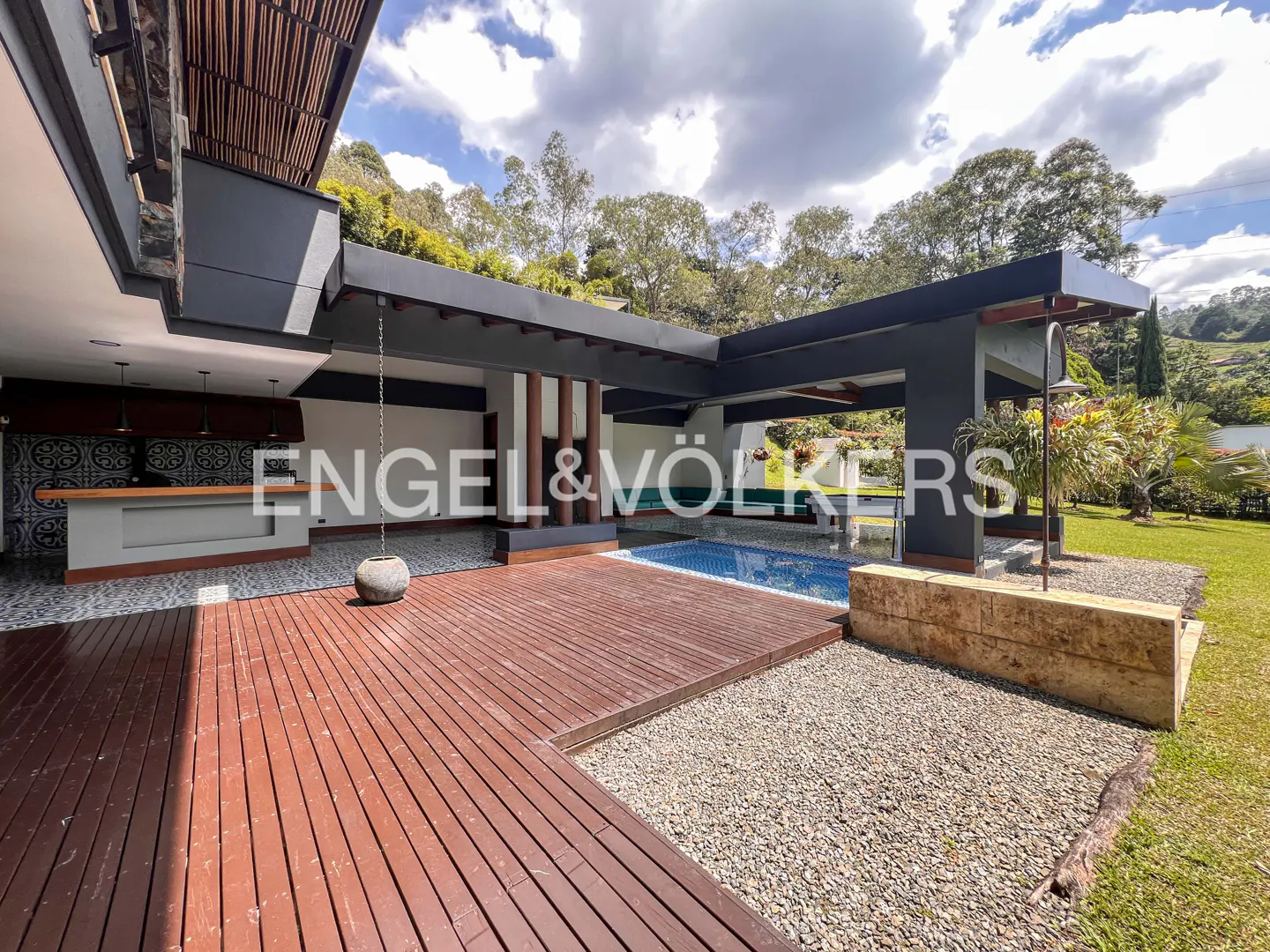 Outdoor patio with a wood deck, pool, and open kitchen area under a dark gray pergola. The Engel & Völkers logo is visible.