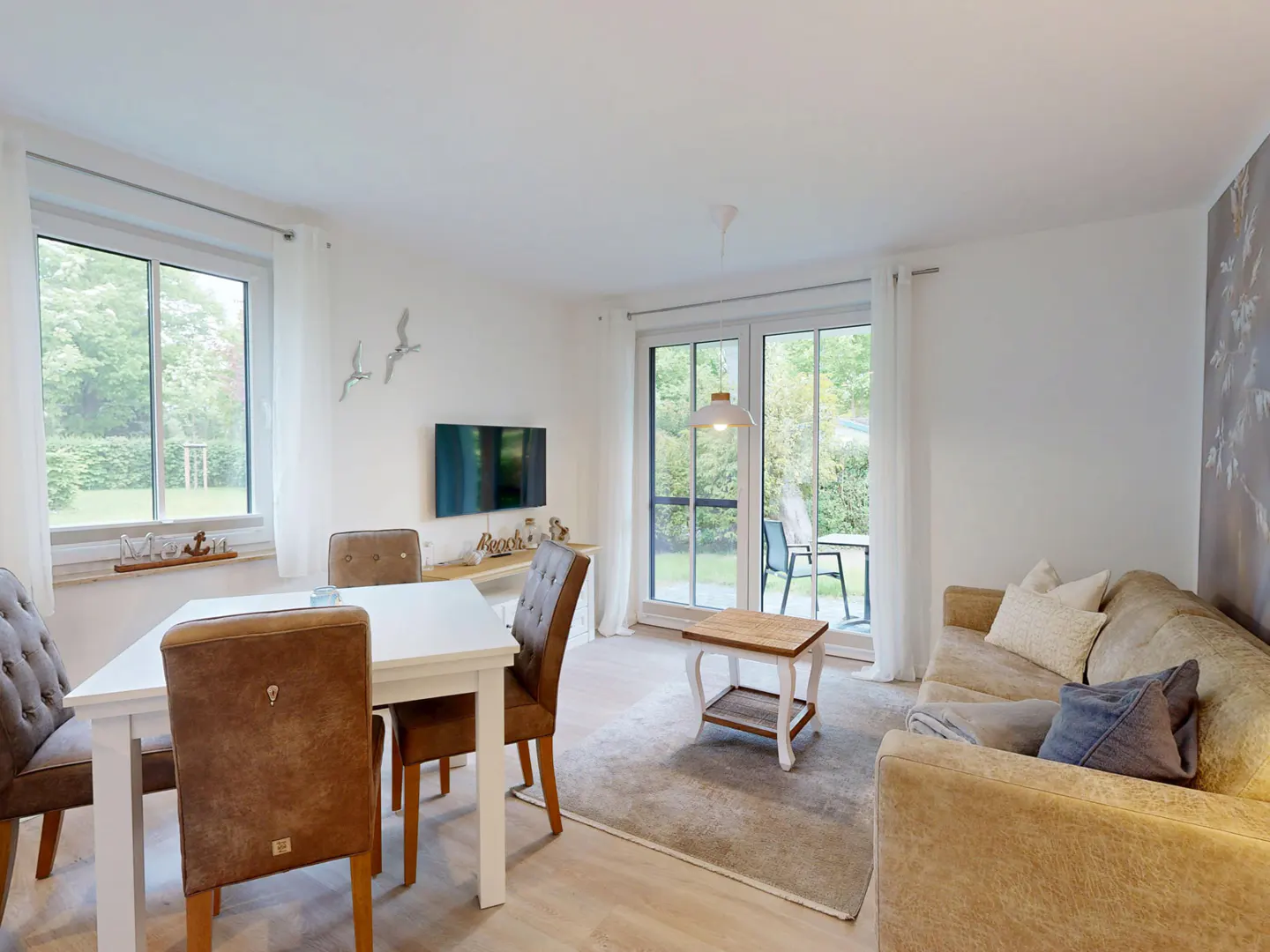 Living room with white walls, a white table with brown chairs, and a beige sofa with blue pillows. A TV is mounted on the wall.