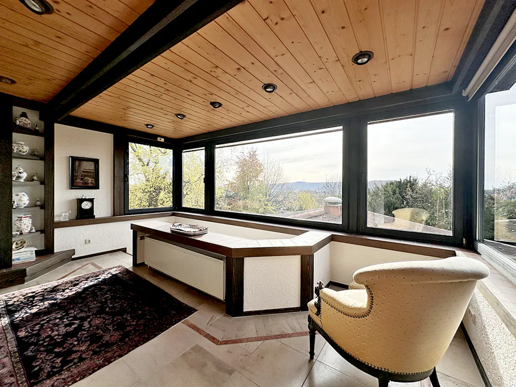 Bright sunroom with wood ceiling, large windows, and a view of trees. A yellow chair sits on a marble floor with a dark rug.