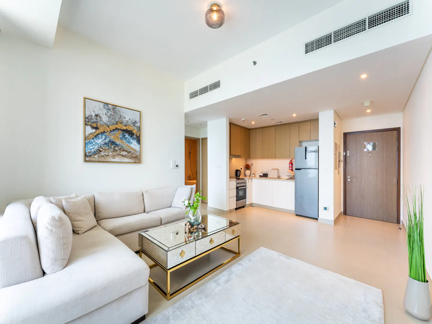 Bright, modern apartment with a beige sectional sofa, mirrored coffee table, and open kitchen. An abstract painting hangs on the white wall.