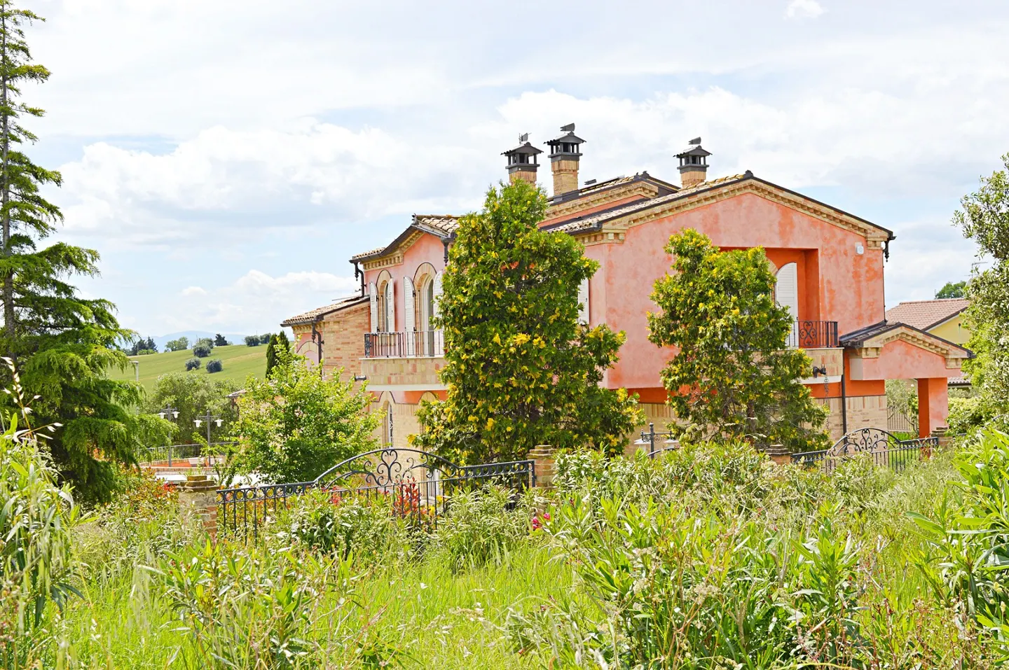 A peach-colored Italian-style villa with three chimneys, surrounded by lush green trees and a wild grassy foreground.